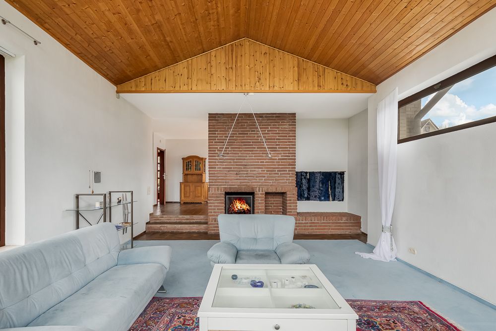 Wohnzimmer mit Holzdecke, Kamin, Sofas und Couchtisch vor großen Fenstern.