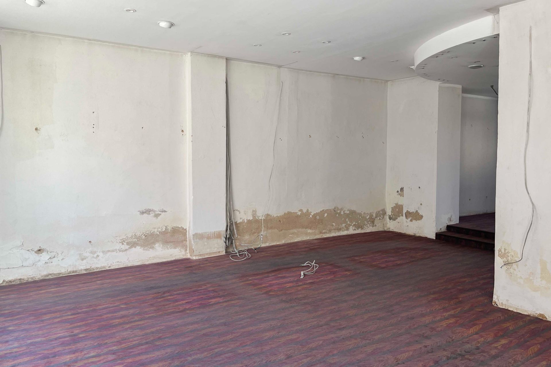 Leerer Raum mit abgenutzten Wänden und dunklem Holzboden, einige Kabel hängen von der Decke.