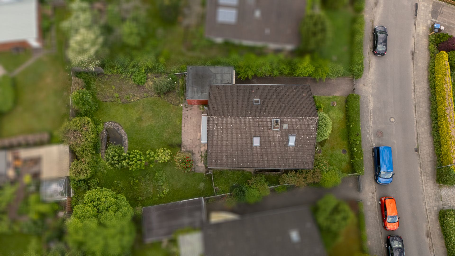 Luftaufnahme eines Einfamilienhauses mit Garten, Auffahrt und parkenden Autos an der Straße.