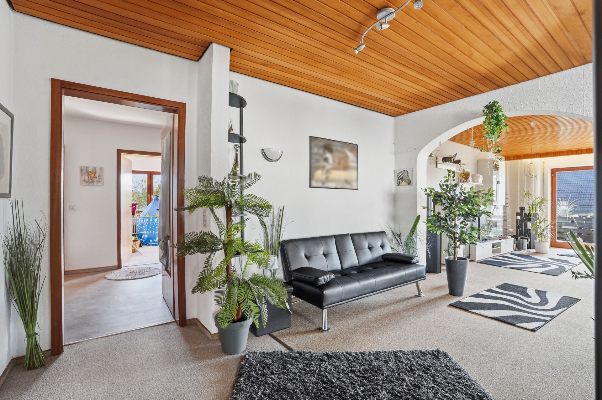 Wohnzimmer mit schwarzem Sofa, Holzdecke, vielen Pflanzen und Zugang zu einem Balkon.