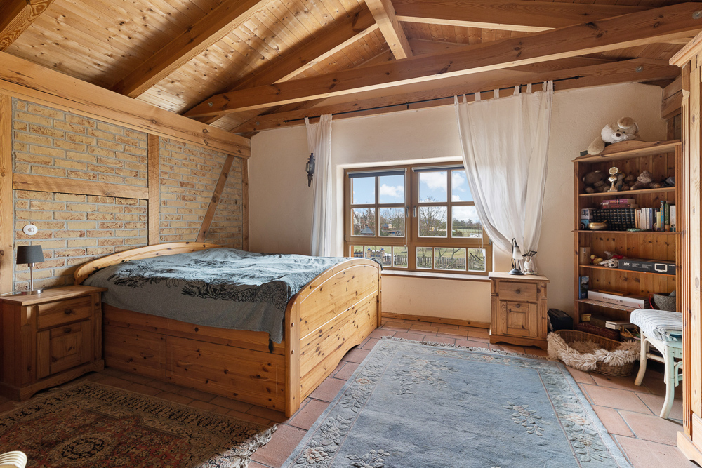 Gemütliches Schlafzimmer mit Holzbett, Bücherregal, Fenster und Holzbalkendecke.