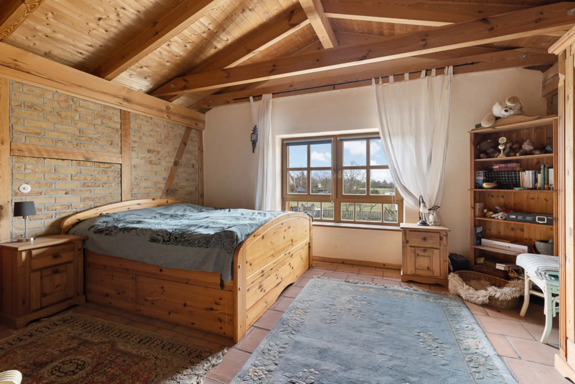 Gemütliches Schlafzimmer mit Holzbett, Bücherregal, Fenster und Holzbalkendecke.