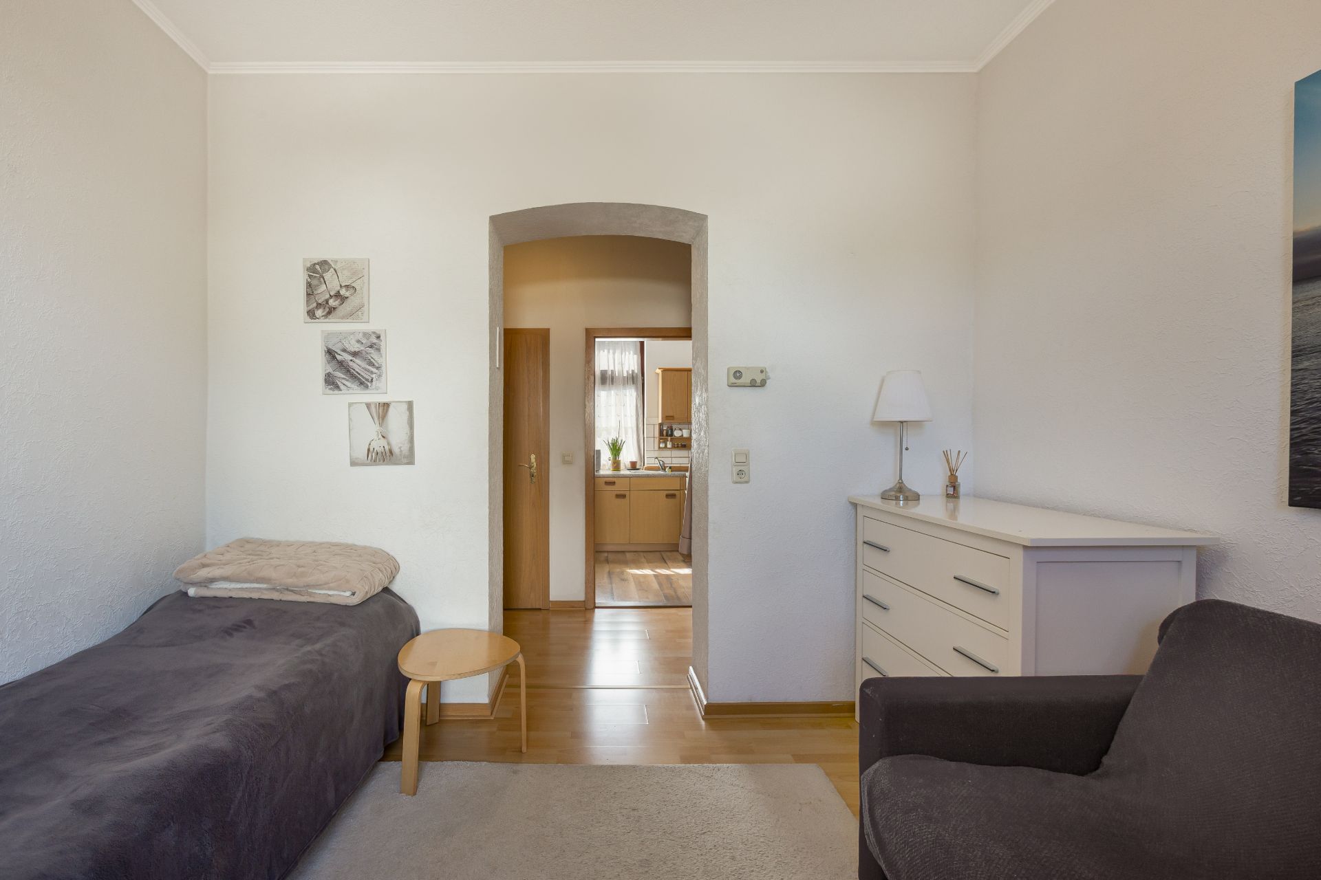 Kleines Zimmer mit Einzelbett, Kommode, Stuhl und Durchgang zu einem weiteren Raum.
