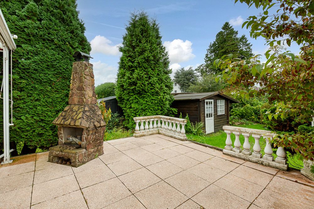 Steinterrasse mit gemauertem Kaminofen, weißen Geländern und Gartenhaus im Hintergrund.