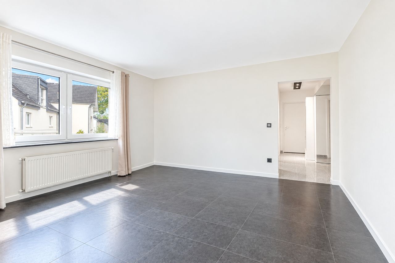 Helles, leeres Zimmer mit großem Fenster, grauem Boden und weiß gestrichenen Wänden.
