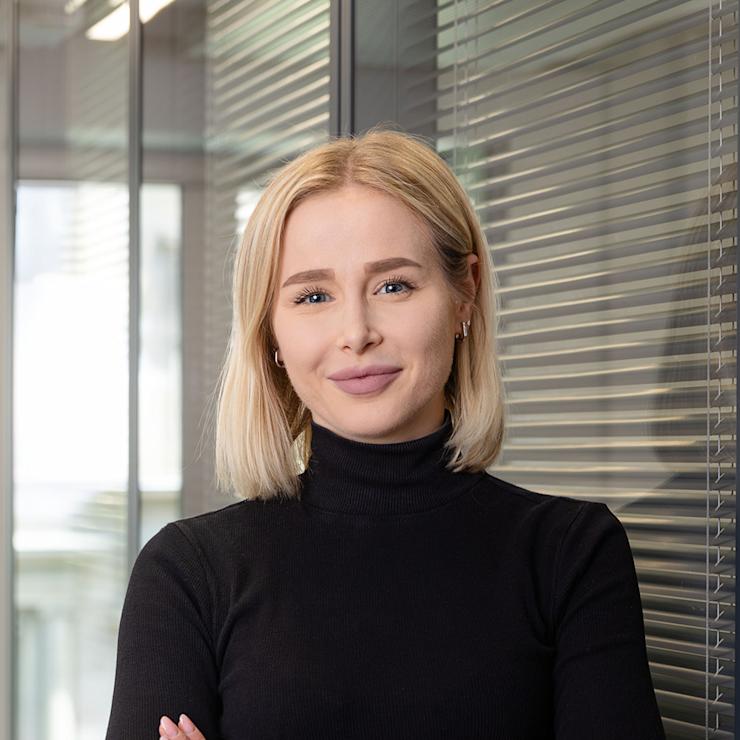 Frau mit blondem Haar und schwarzem Rollkragenpullover steht vor Glaswand.