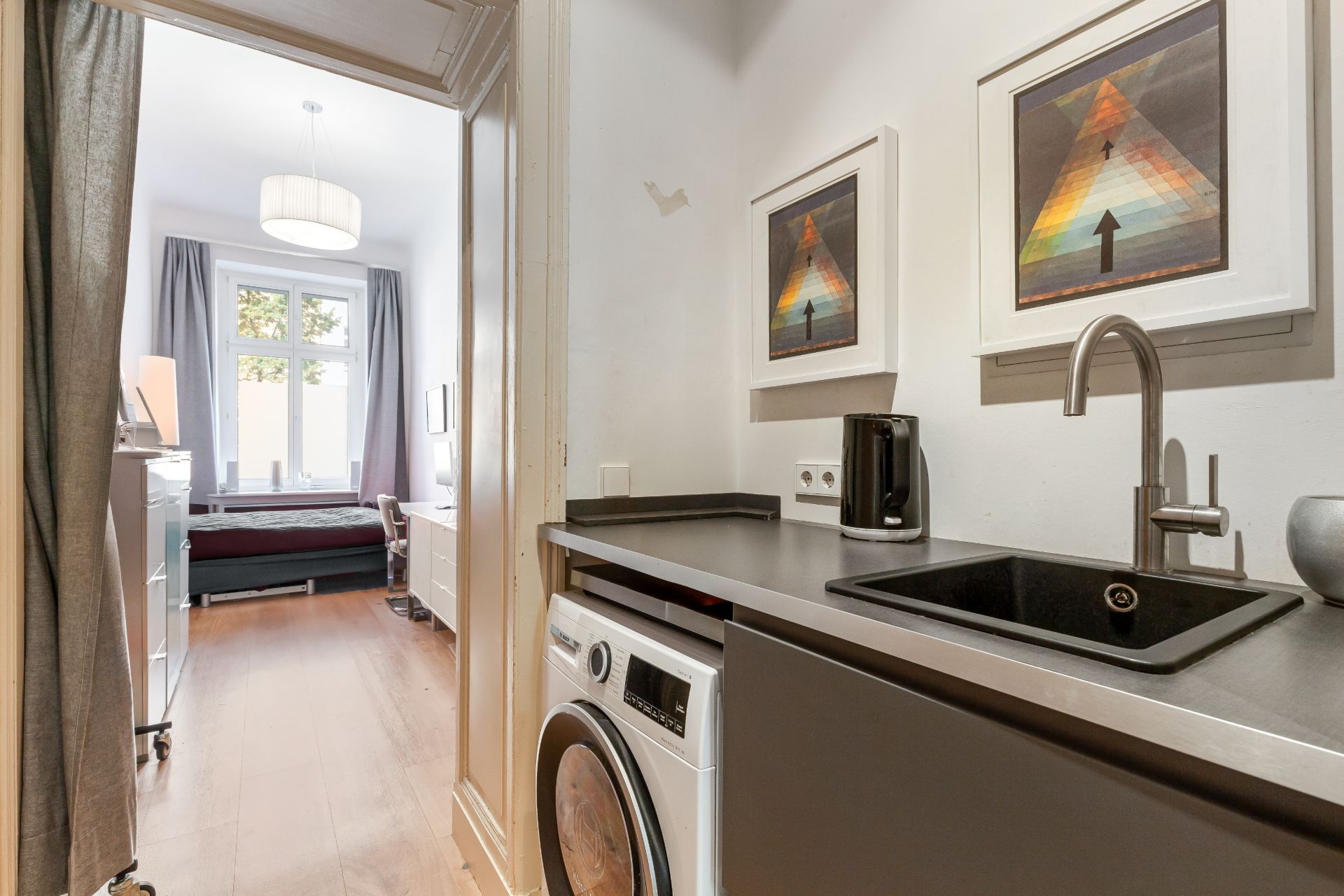 Kleine Küche mit Waschmaschine, Spüle und zwei Bildern, dahinter Schlafzimmer mit großem Fenster.