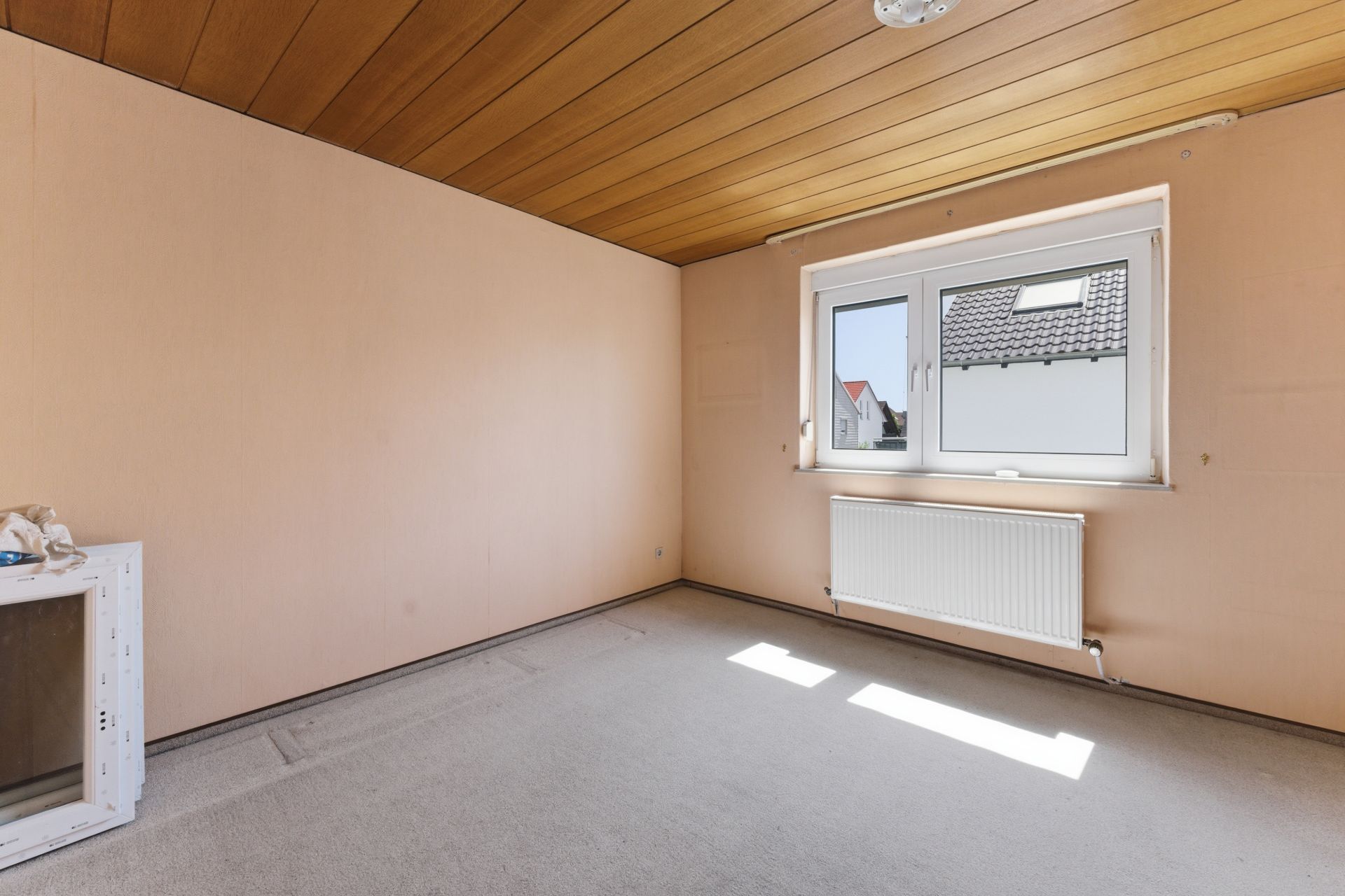 Leeres Zimmer mit Teppichboden, Holzdecke, Fenster und Heizkörper an der Wand.