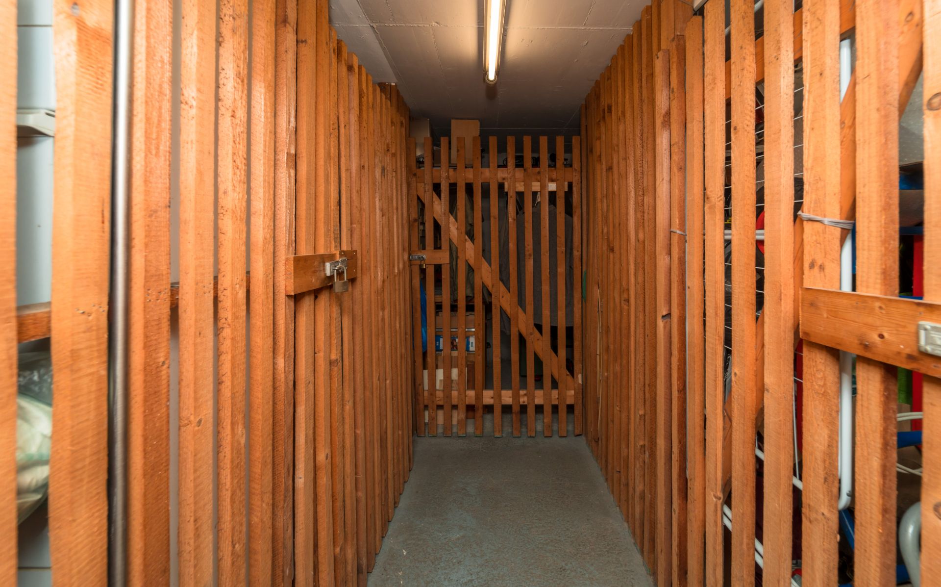 Schmaler Kellerraum mit Holzgitterwänden und Betonboden, beleuchtet durch eine Deckenlampe.