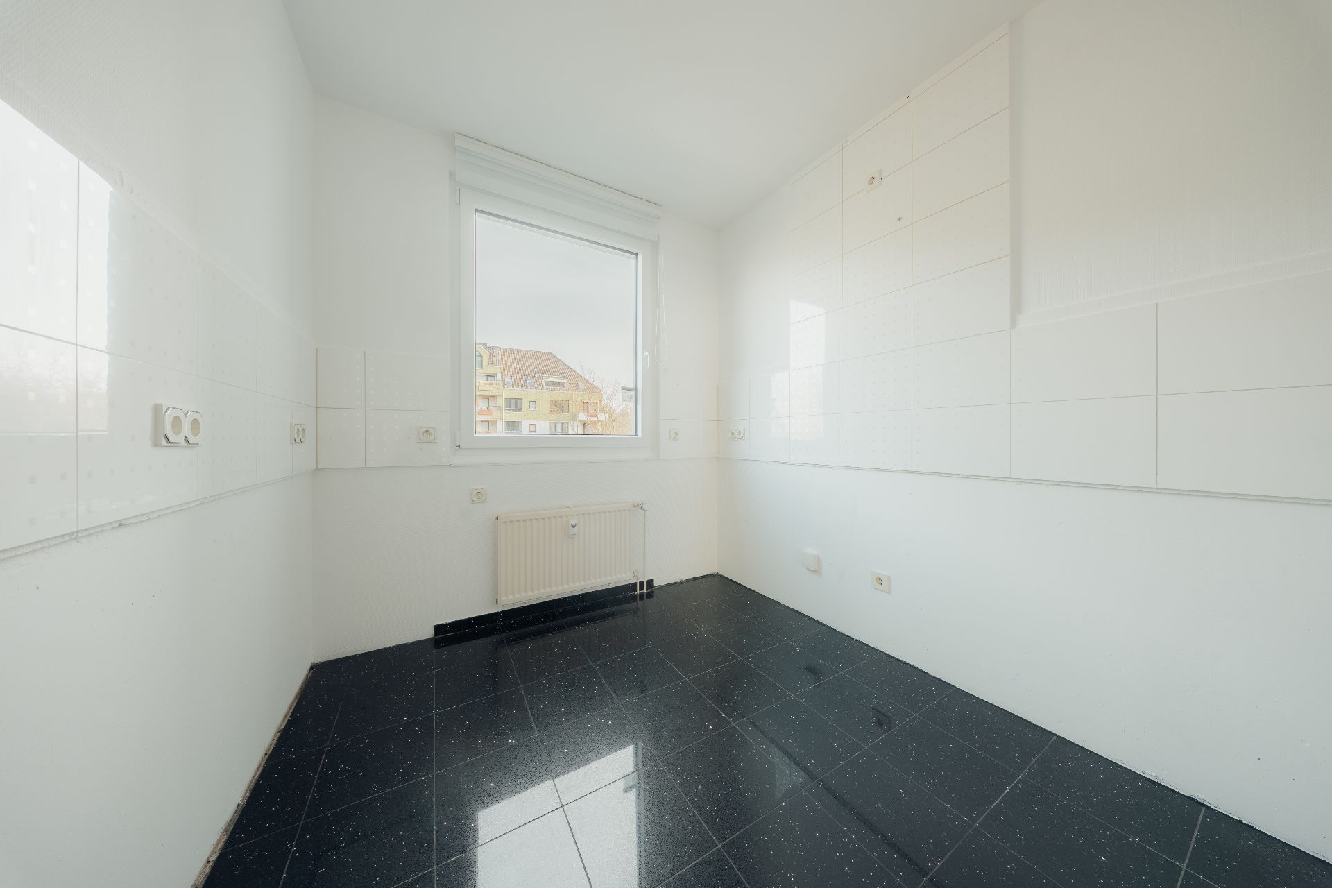 Leerer, heller Raum mit schwarzem Fliesenboden und einem Fenster, Blick nach draußen.