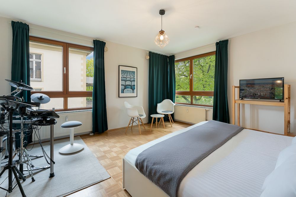 Helles Schlafzimmer mit Doppelbett, elektronischem Schlagzeug, zwei Stühlen, Fernseher und großen Fenstern.