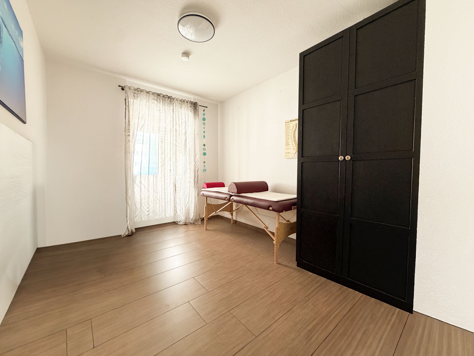 Helles Zimmer mit Holzfußboden, großem schwarzen Schrank und Liege am Fenster.