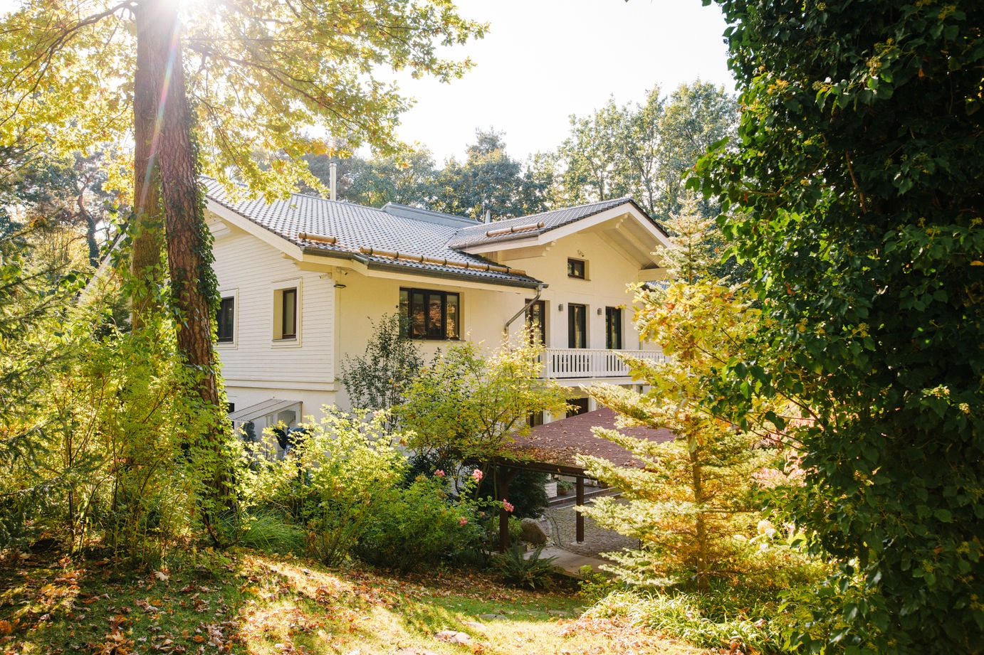 Zweistöckiges weißes Haus mit Balkon, umgeben von Bäumen und viel Grün.