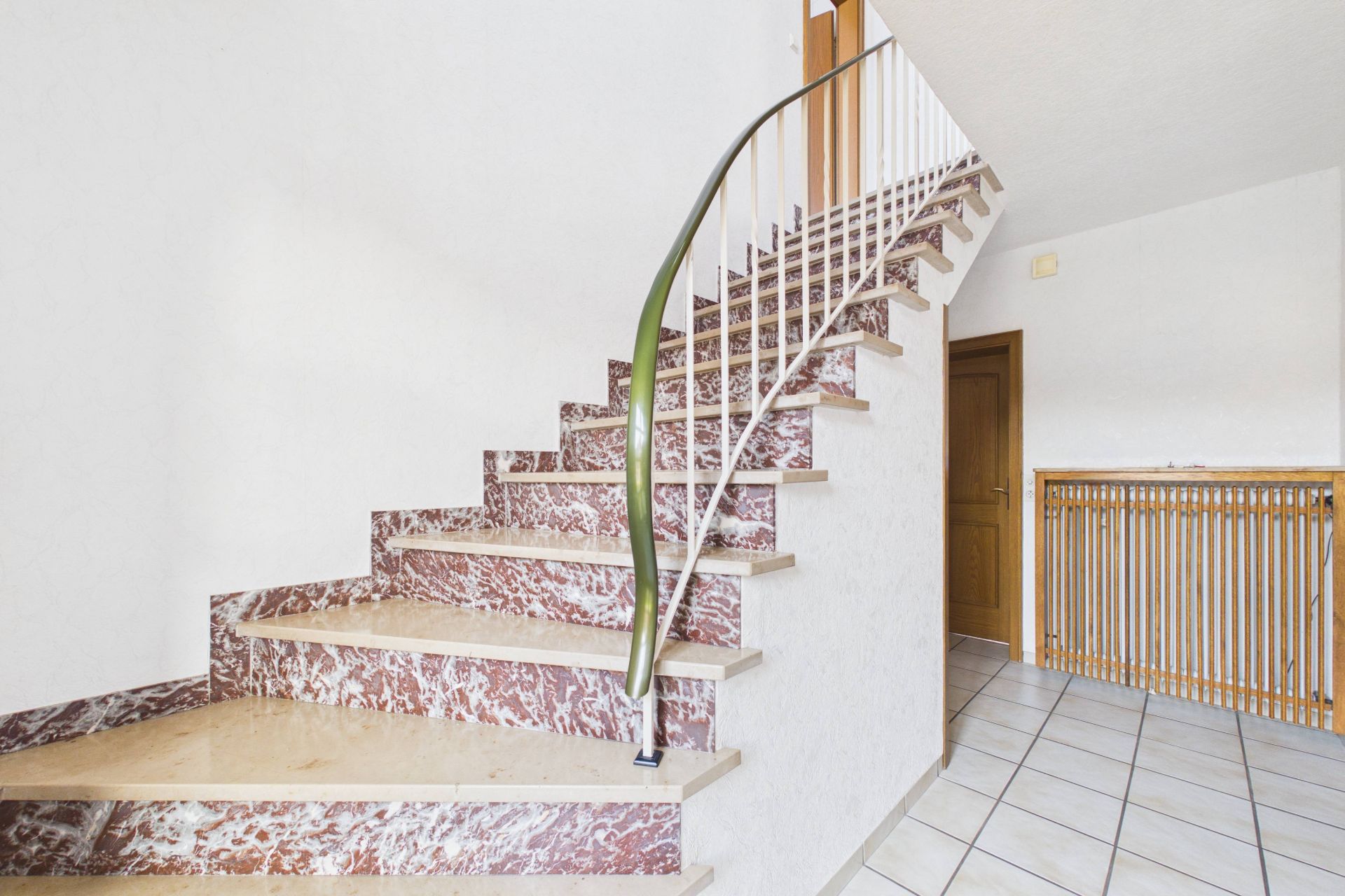 Treppe mit rot-weißem Marmormuster und grünem Handlauf in einem Flur mit Fliesenboden.