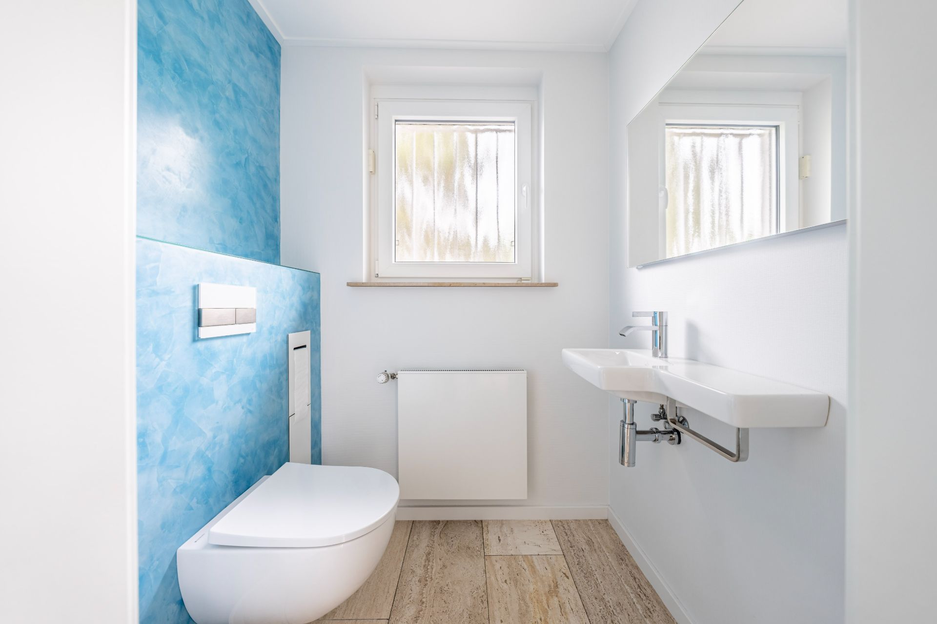 Helles Badezimmer mit wandhängendem WC, Waschbecken, Fenster und blau gestrichener Wand.