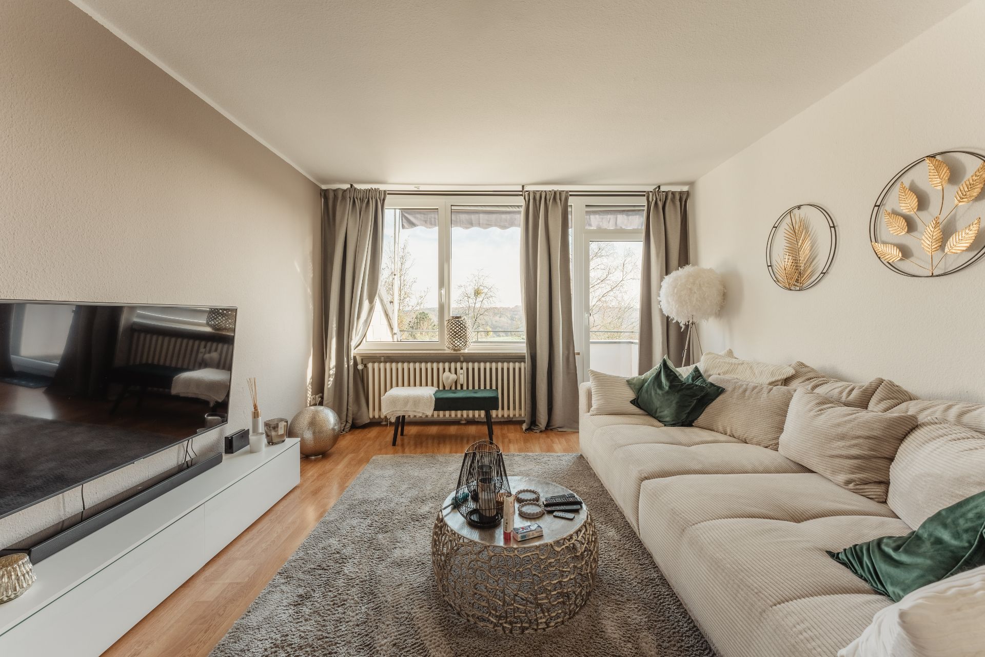 Helles Wohnzimmer mit beigem Sofa, rundem Tisch, Fernseher und großen Fenstern mit Vorhängen.