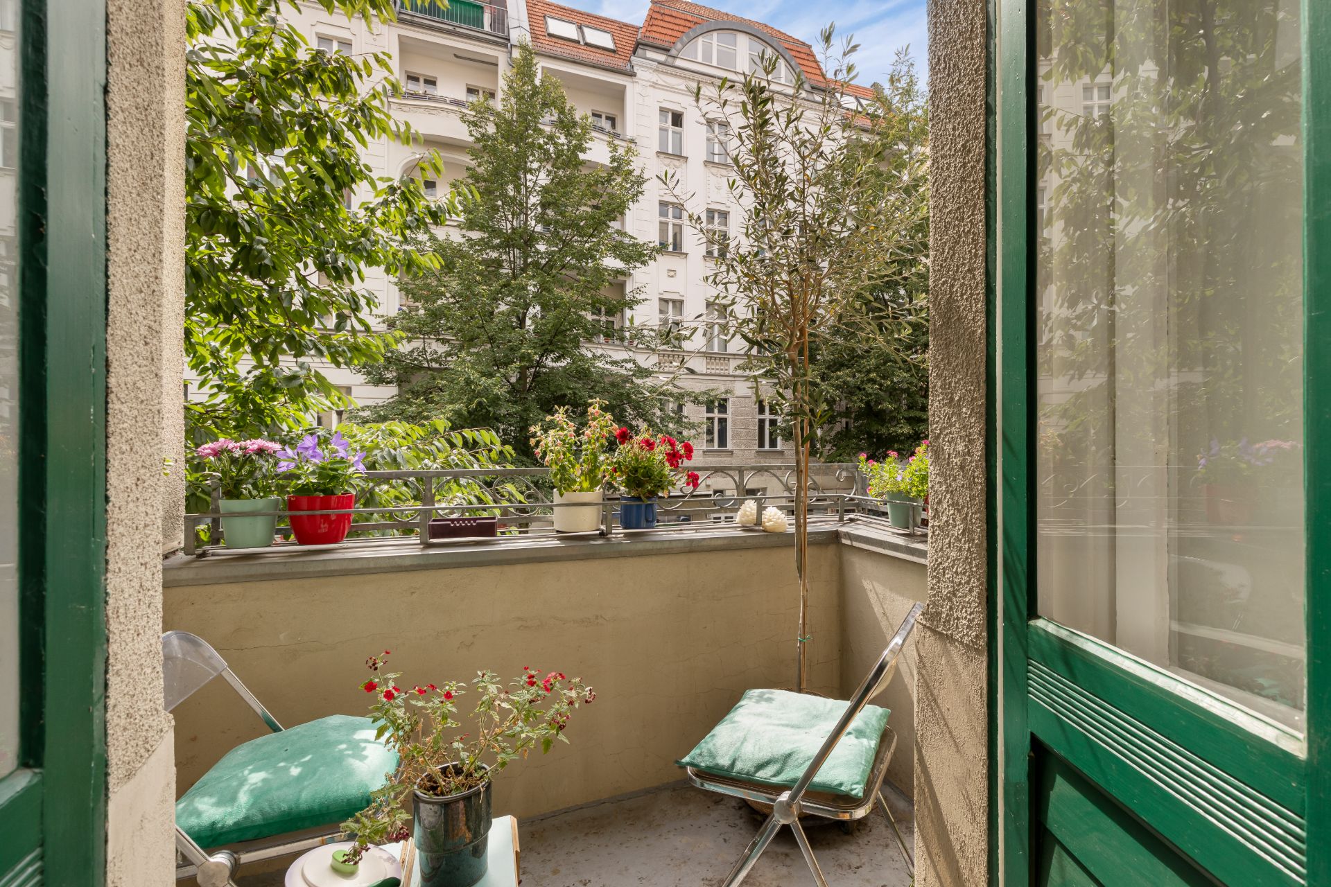 Kleiner Balkon mit Stühlen, grünen Kissen und vielen Blumentöpfen, Blick auf Nachbargebäude.