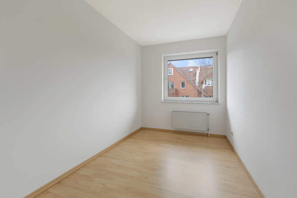 Leerer Raum mit hellen Wänden, Holzfußboden und Fenster mit Blick auf ein rotes Gebäude.