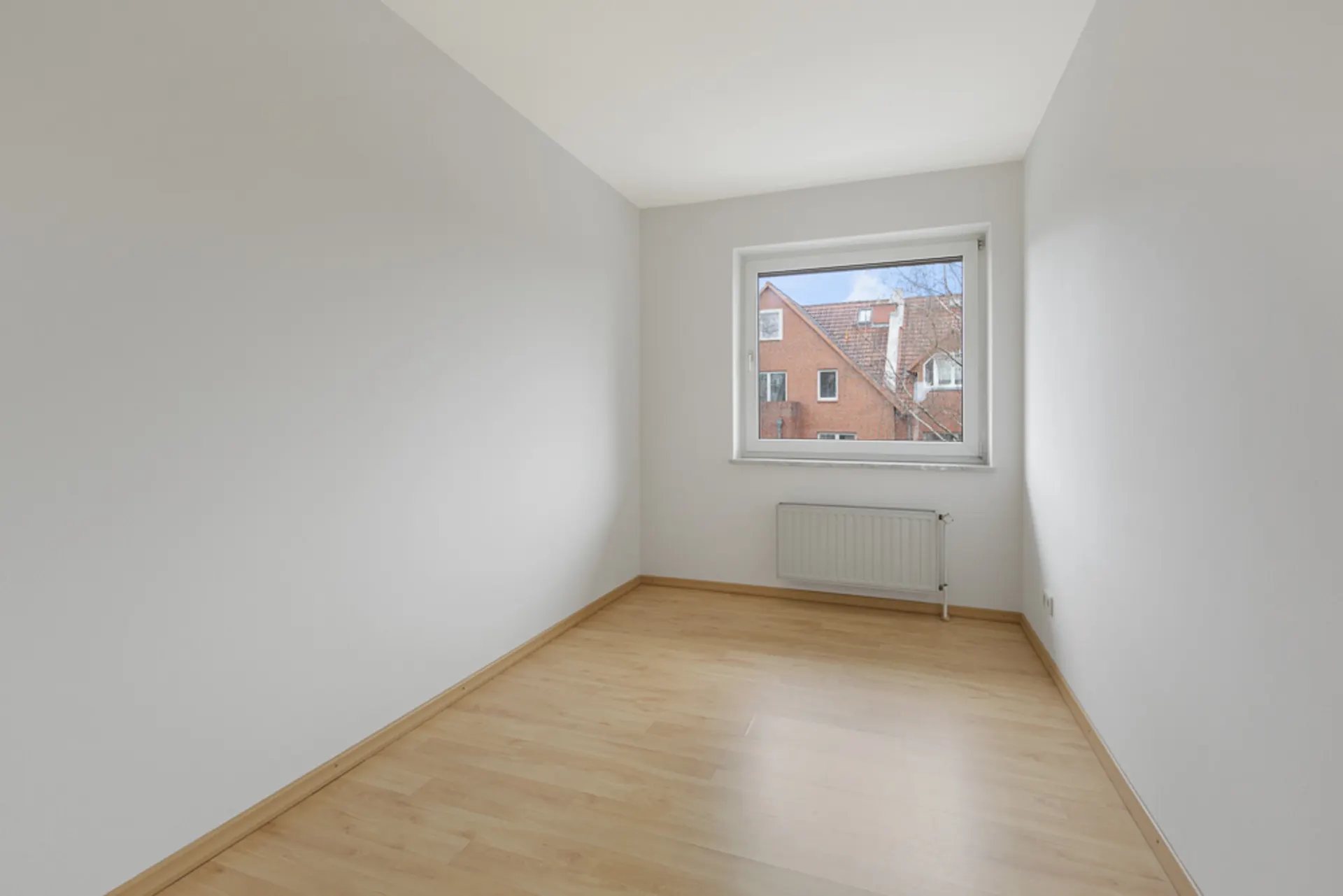 Leerer Raum mit hellen Wänden, Holzfußboden und Fenster mit Blick auf ein rotes Gebäude.