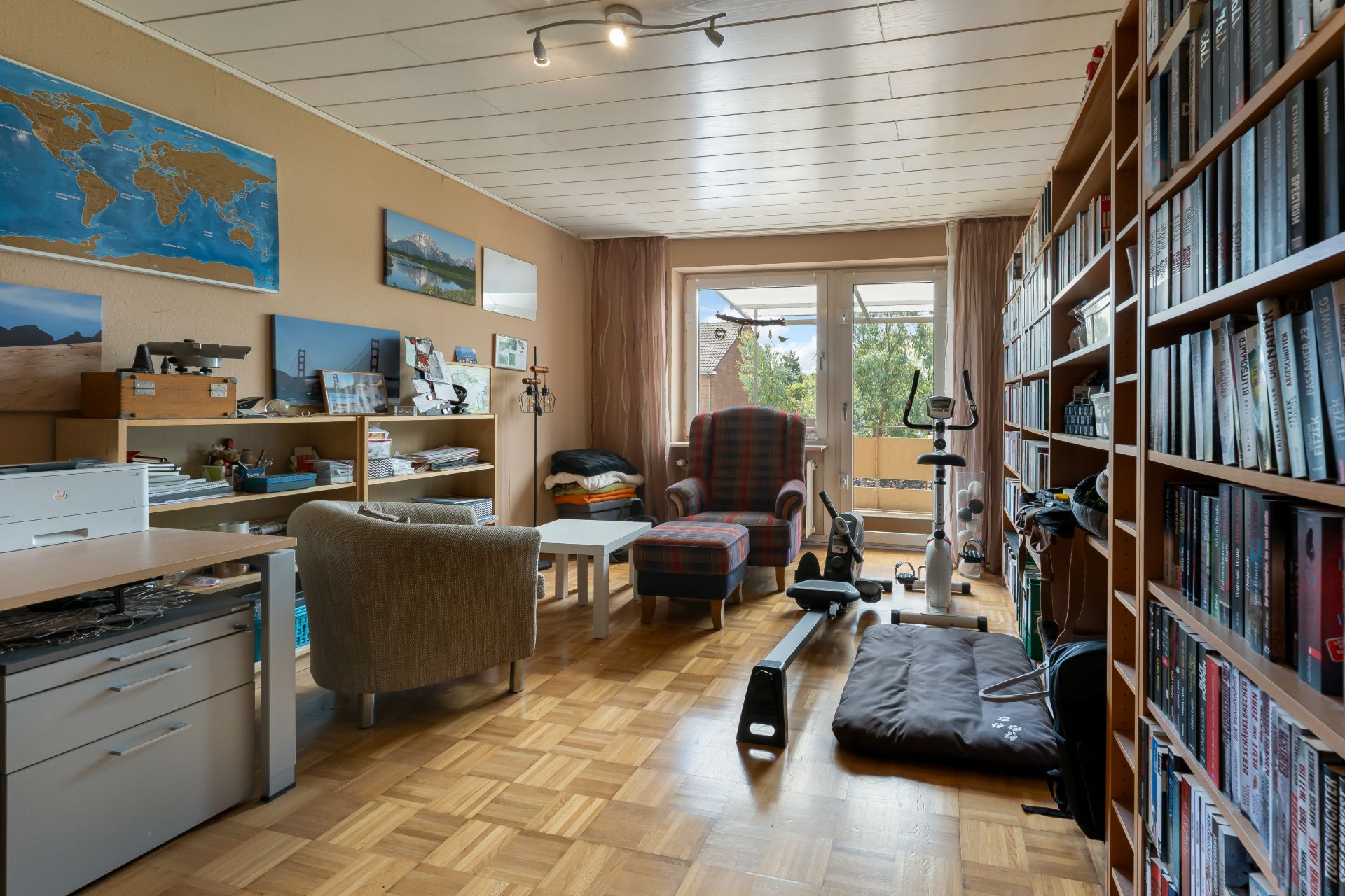 Helles Zimmer mit Bücherregal, Schreibtisch, Sesseln, Fitnessgeräten und Wandbildern.