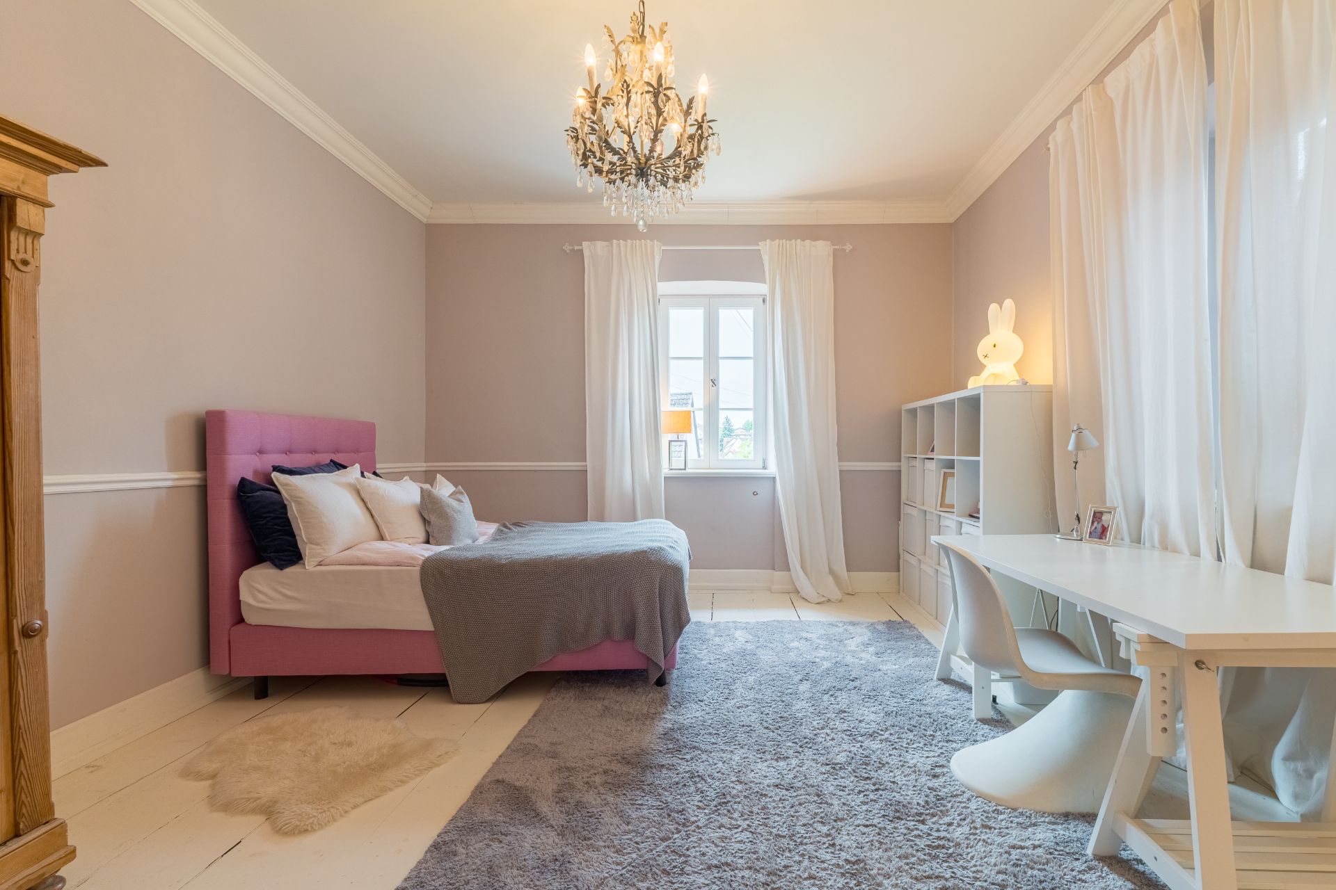 Helles Schlafzimmer mit rosa Bett, Schreibtisch, Regal und Kronleuchter.