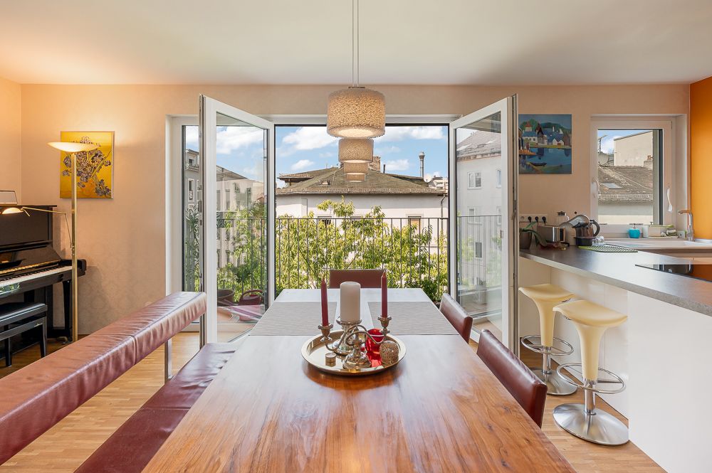 Essbereich mit Holztisch, offener Küche, Klavier und großer Fensterfront mit Blick nach draußen.