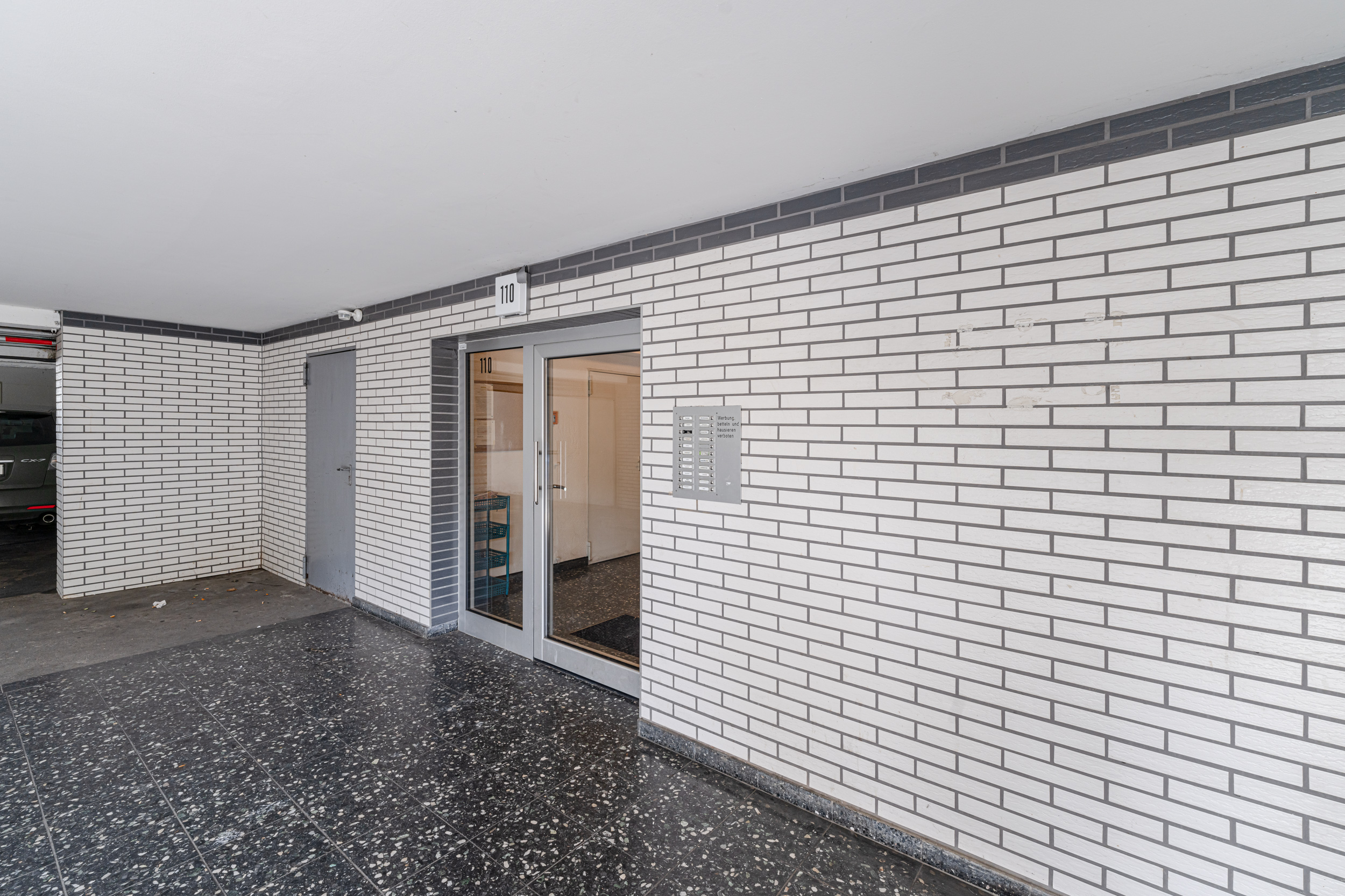 Eingang eines Wohngebäudes mit Glastür, Klingelanlage und weißer Klinkerwand.