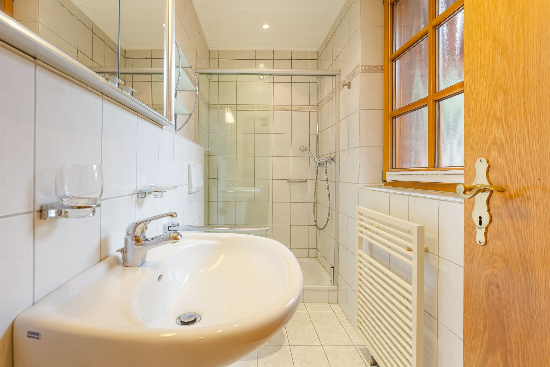 Helles Badezimmer mit Waschbecken, bodengleicher Dusche und Fenster mit Holzrahmen.