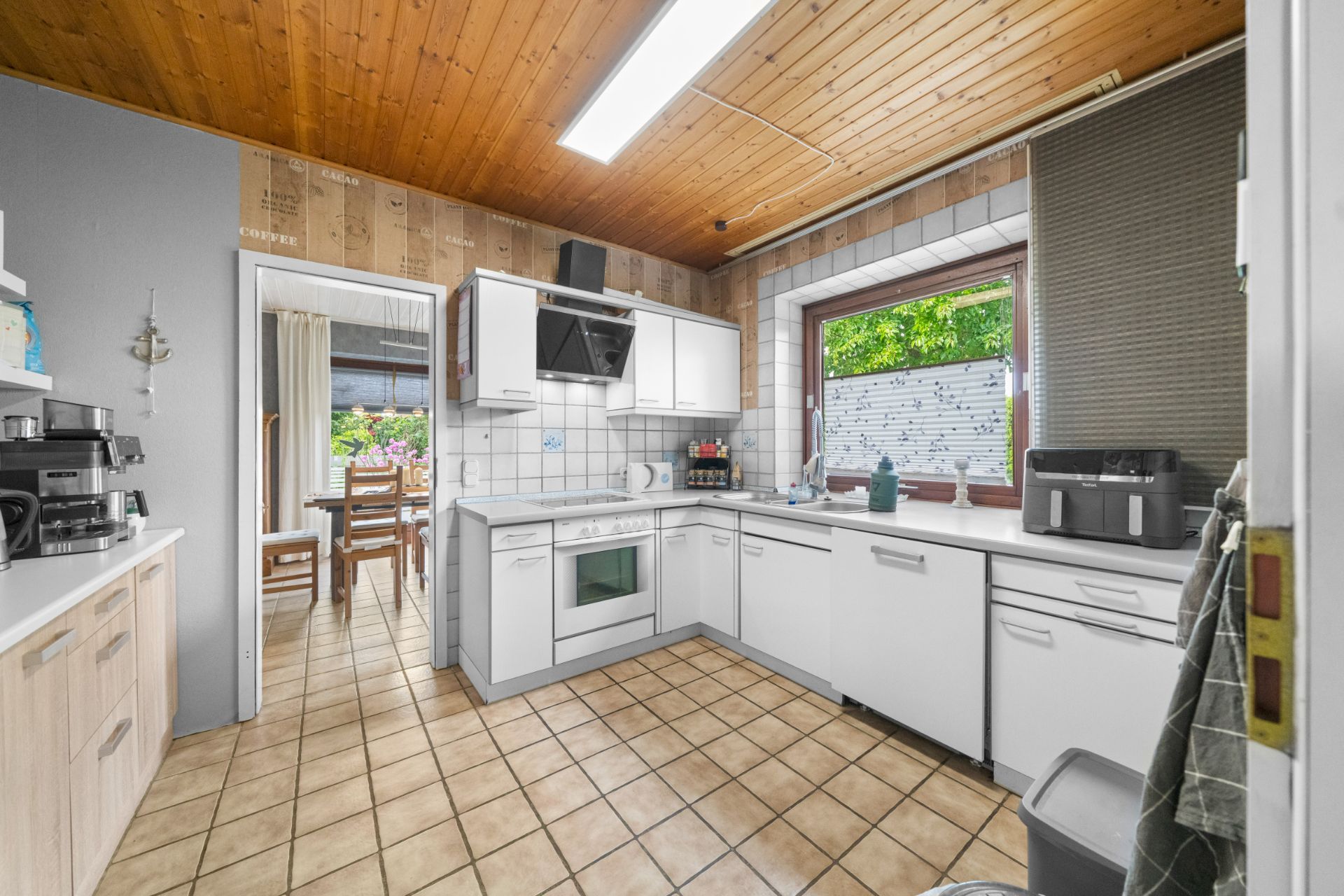 Helle Küche mit weißen Schränken, großem Fenster und Fliesenboden, Durchgang zum Esszimmer.