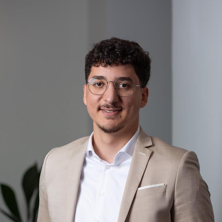 Junger Mann mit Brille und beigem Sakko steht vor einer Pflanze in einem modernen Raum.