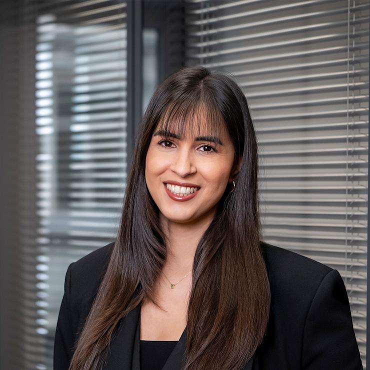 Frau mit langen dunklen Haaren und schwarzem Blazer steht vor einem Fenster mit Jalousien.