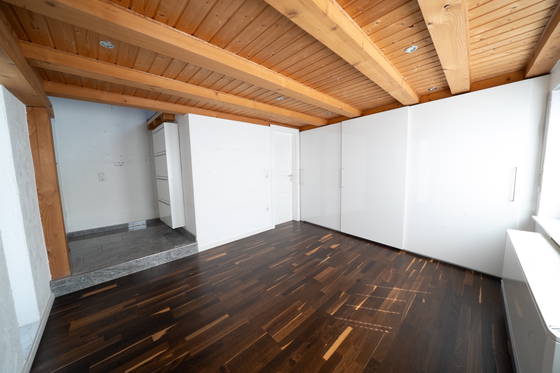 Leeres Zimmer mit dunklem Holzboden, weißen Schränken und Holzdecke.