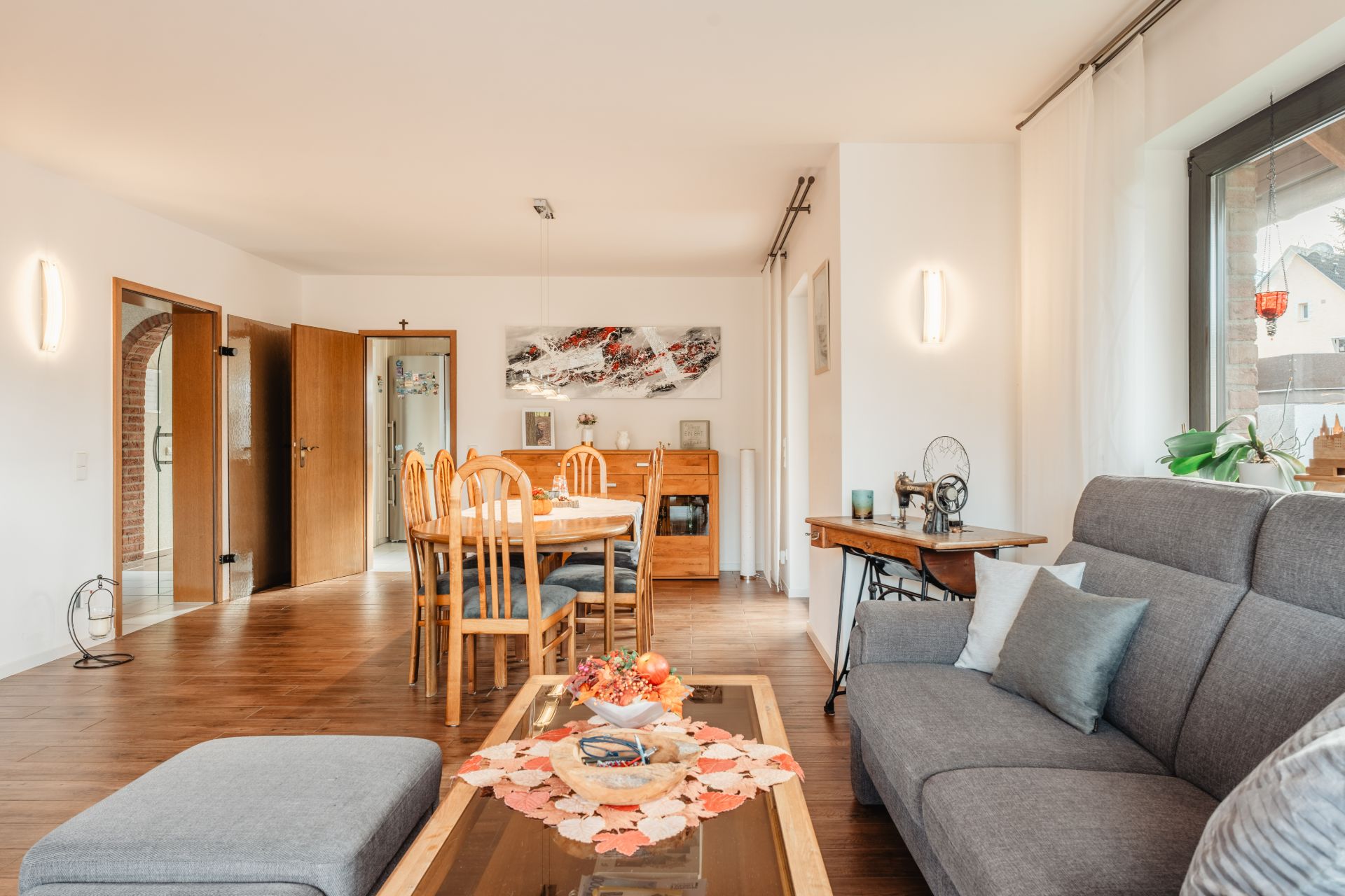 Helles Wohnzimmer mit grauem Sofa, Esstisch, Holzmöbeln und großer Fensterfront.