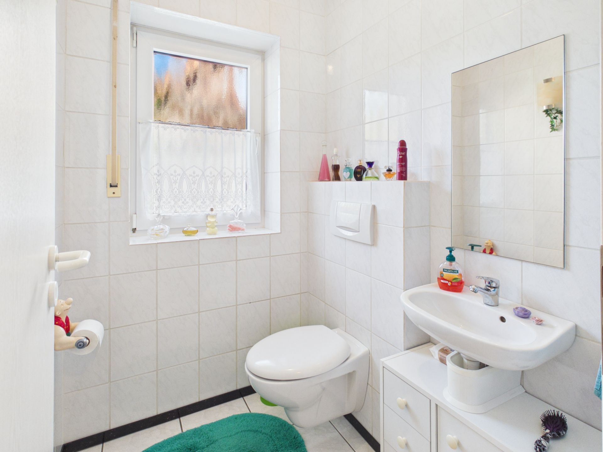 Helles Badezimmer mit WC, Waschbecken, Spiegel und Fenster mit Vorhang.