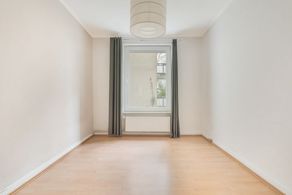Leerer Raum mit hellem Boden, Fenster und dunklen Vorhängen, weiße Wände und Deckenlampe.