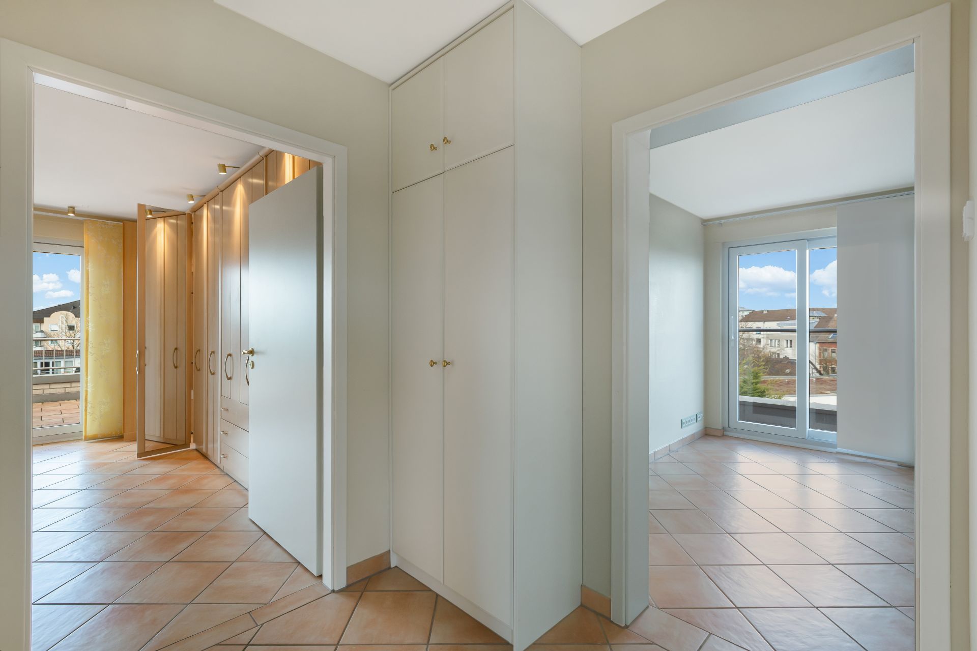 Flur mit Einbauschrank, Türen zu zwei hellen Räumen mit Fliesenboden und großen Fenstern.