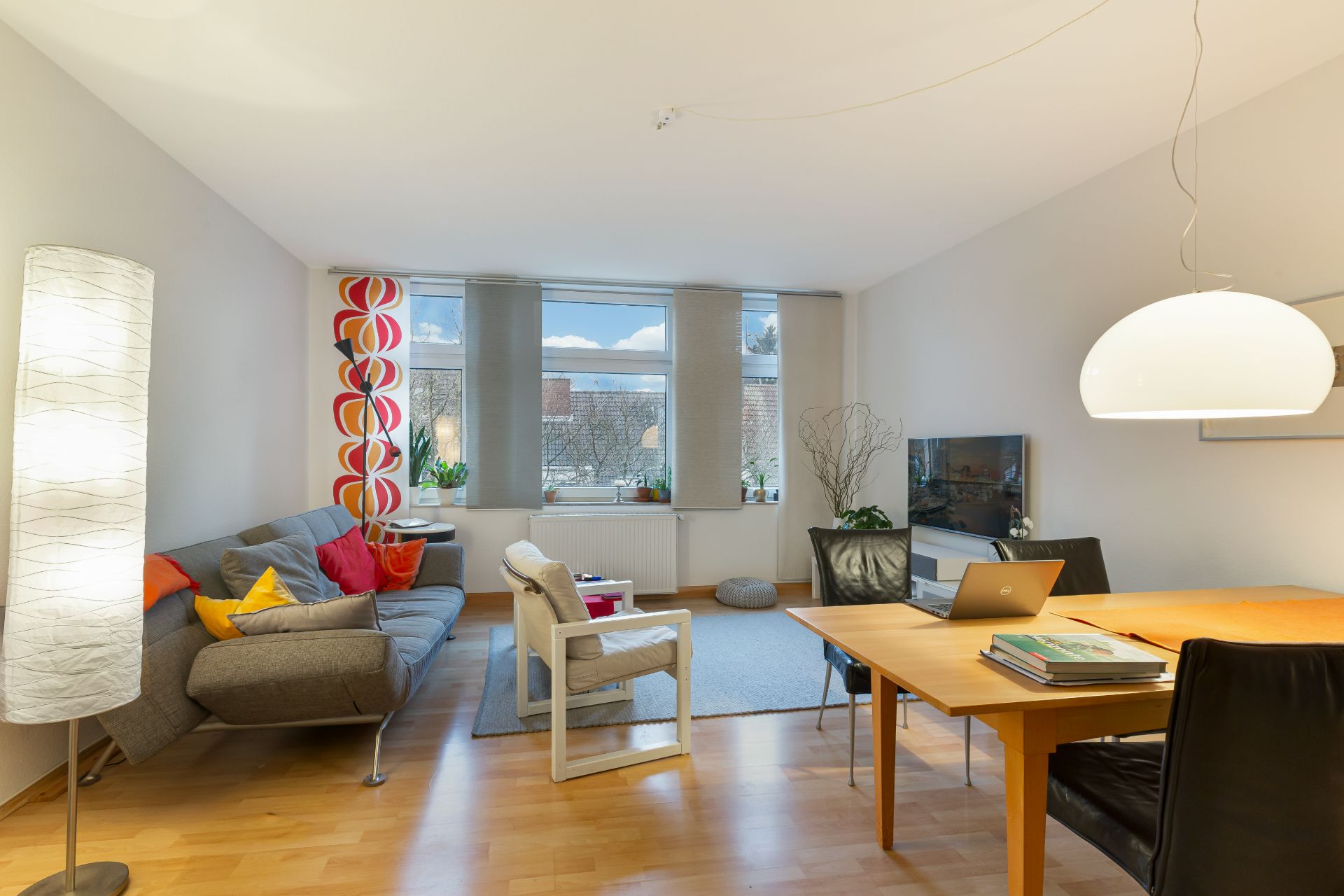 Helles Wohnzimmer mit Sofa, Sessel, Esstisch, großen Fenstern und Pflanzen.