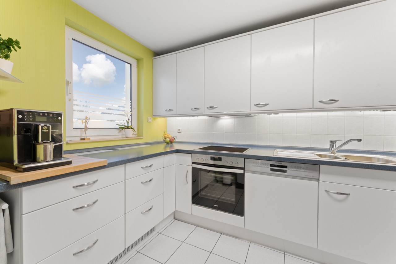 Moderne Küche mit weißen Schränken, Kaffeemaschine, Herd, Spülmaschine und Fenster.