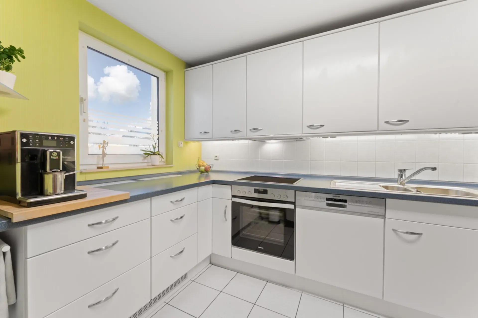 Moderne Küche mit weißen Schränken, Kaffeemaschine, Herd, Spülmaschine und Fenster.
