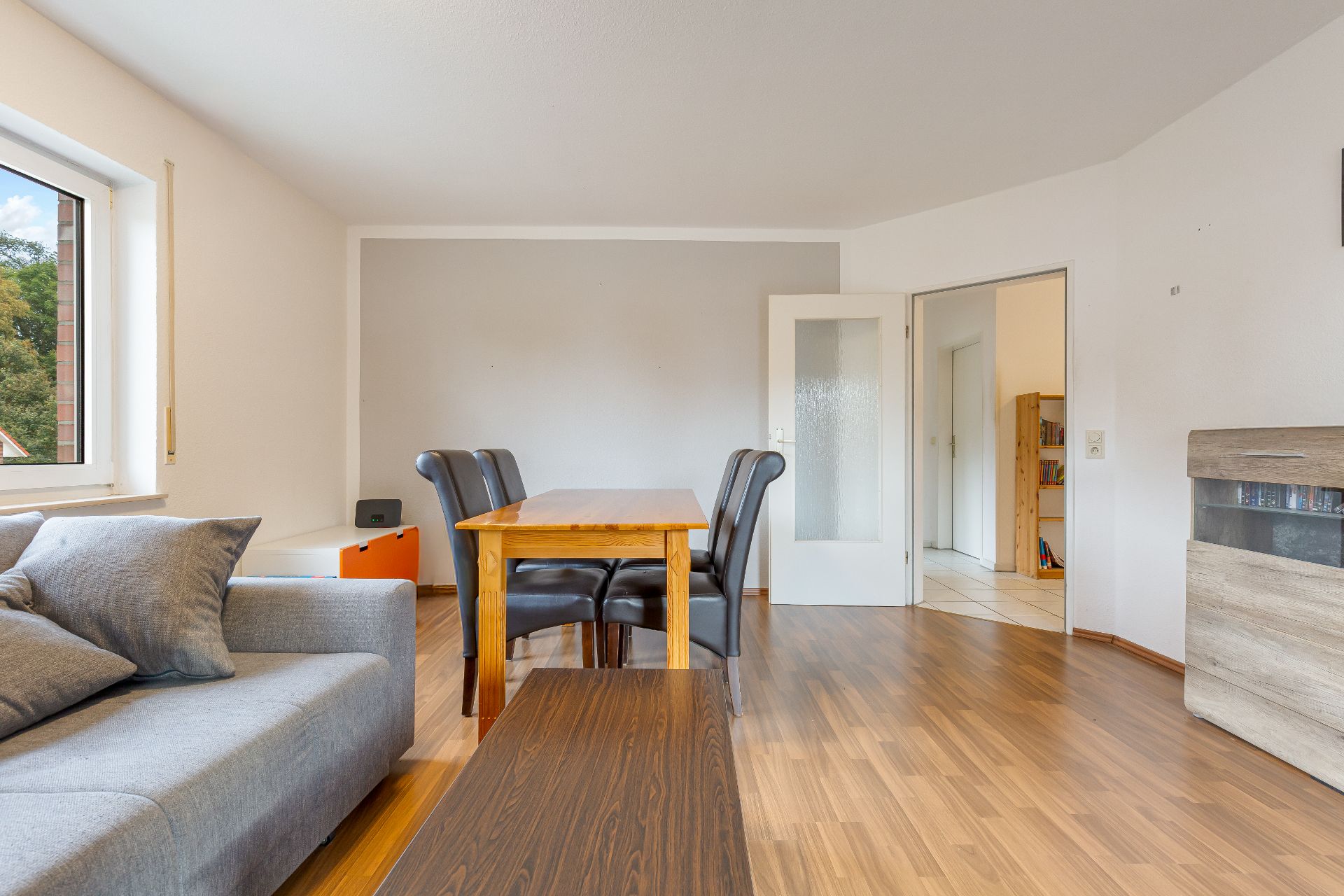 Wohnzimmer mit Holzboden, grauem Sofa, Esstisch und vier Stühlen, Fenster links.