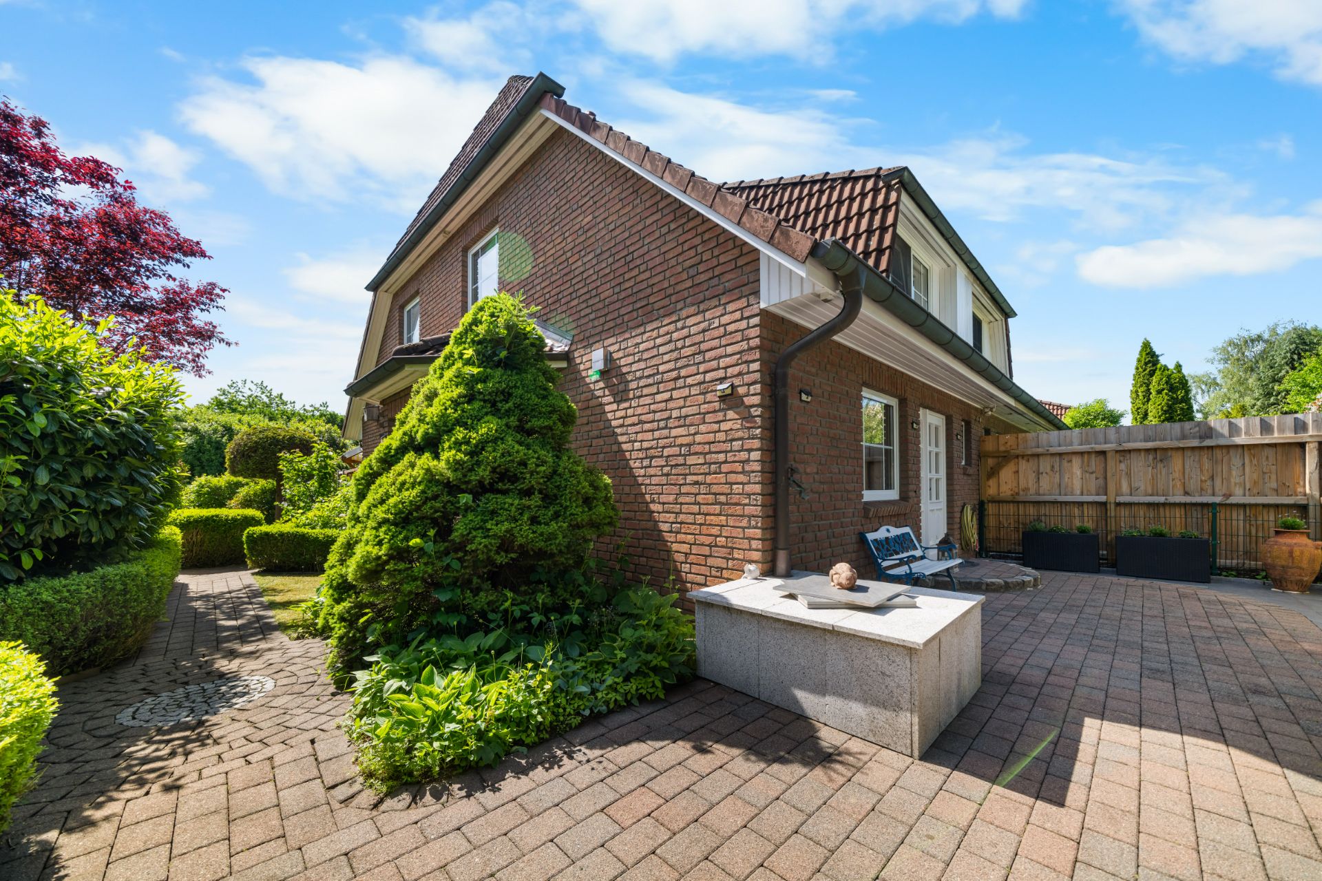 Backsteinhaus mit Terrasse, Gartenweg und gepflegten Sträuchern unter blauem Himmel.