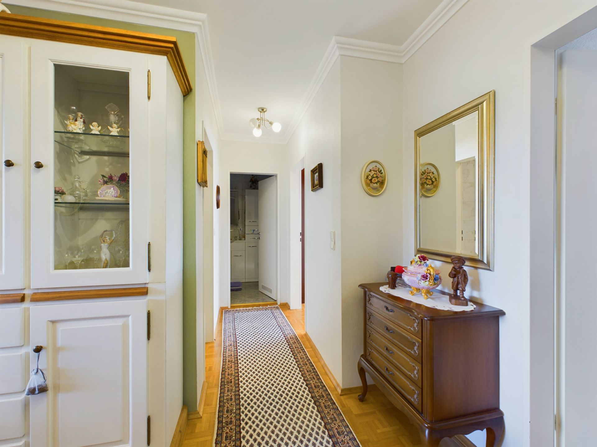 Heller Flur mit Teppichläufer, Vitrine, Spiegel und Kommode mit Dekorationen.
