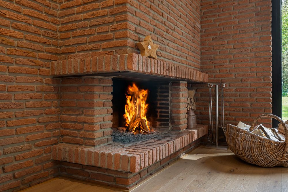 Gemauerter Kamin mit brennendem Feuer und Holzkorb in einer Ecke eines Zimmers.