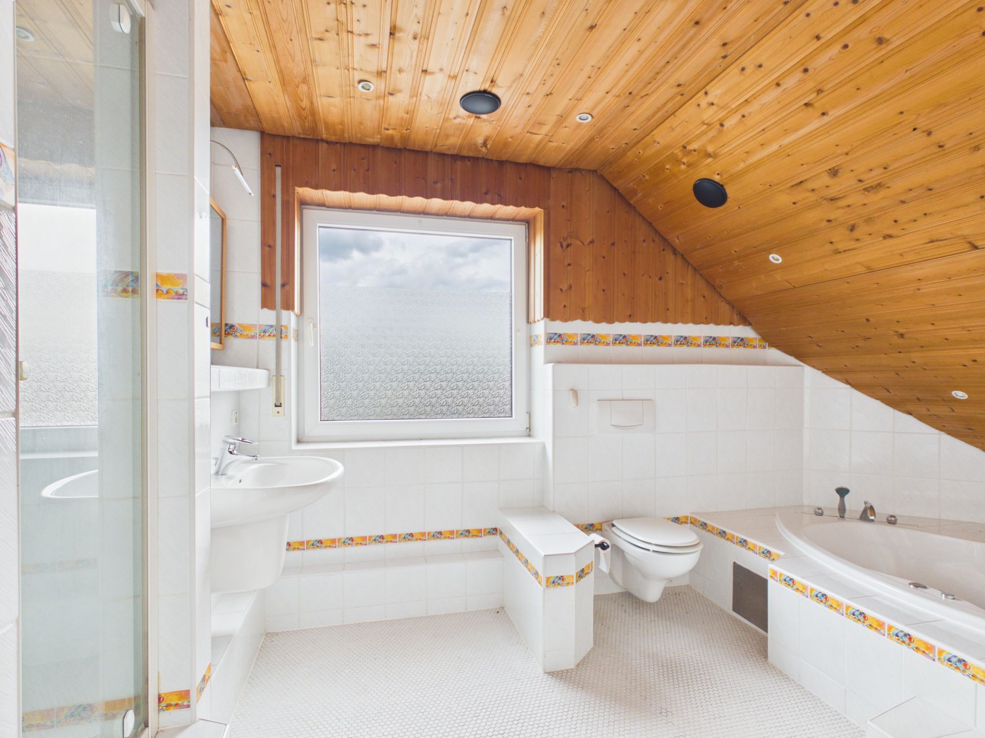 Helles Badezimmer mit Dachschräge, Holzdecke, Badewanne, Toilette, Waschbecken und Fenster.