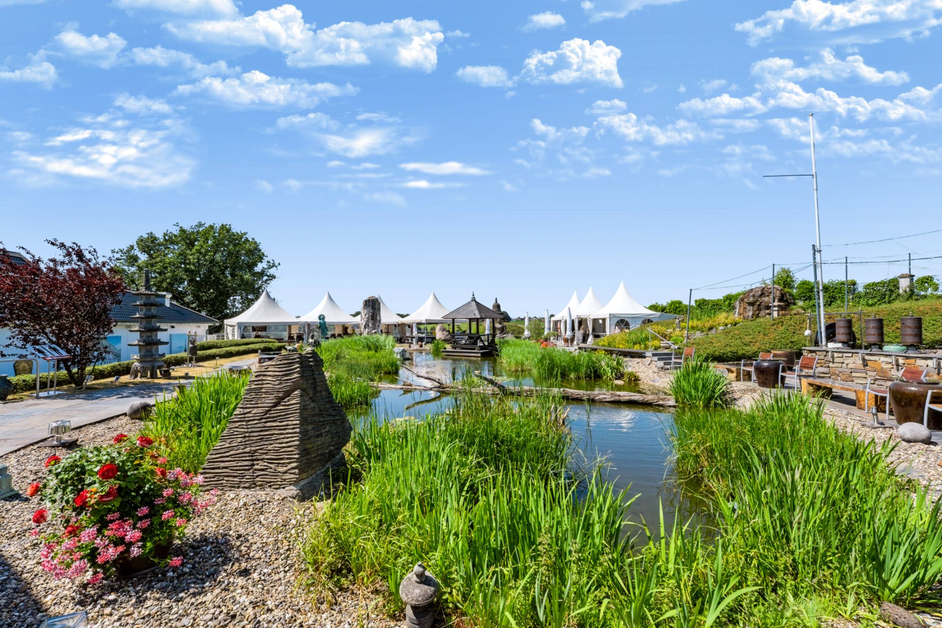 Großer Gartenteich mit Steinen, Blumen und weißen Pavillons im Hintergrund.