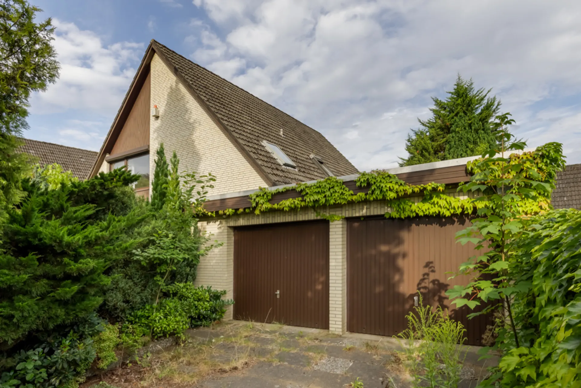 Haus mit Satteldach, zwei braunen Garagentoren und viel Bewuchs davor.