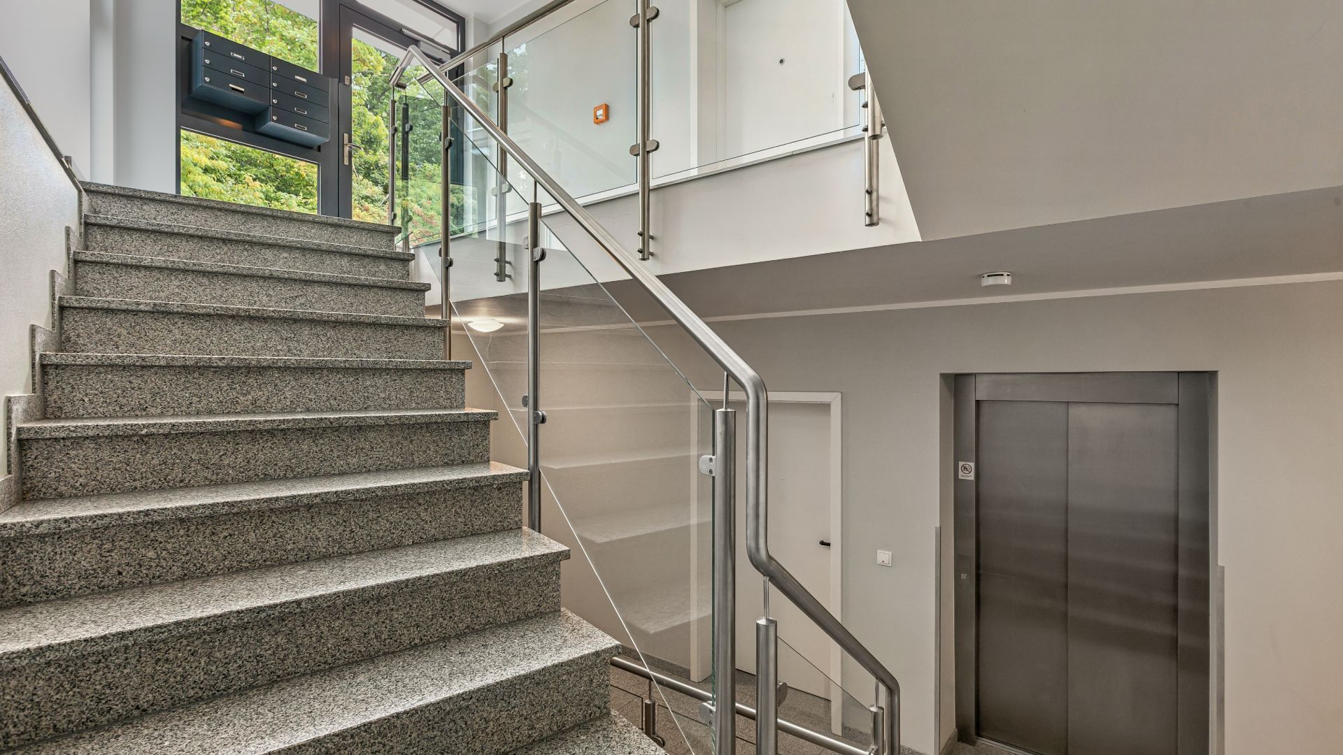 Treppenhaus mit Glasgeländer und Aufzug in einem Wohngebäude.
