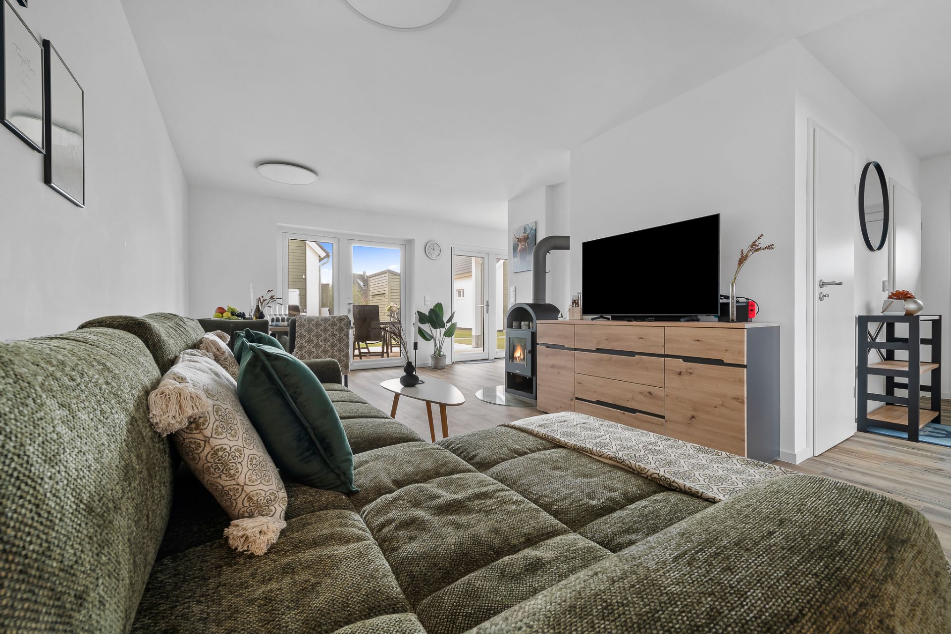Wohnzimmer mit grünem Sofa, Fernseher, Kamin und Zugang zur Terrasse, Essbereich im Hintergrund.
