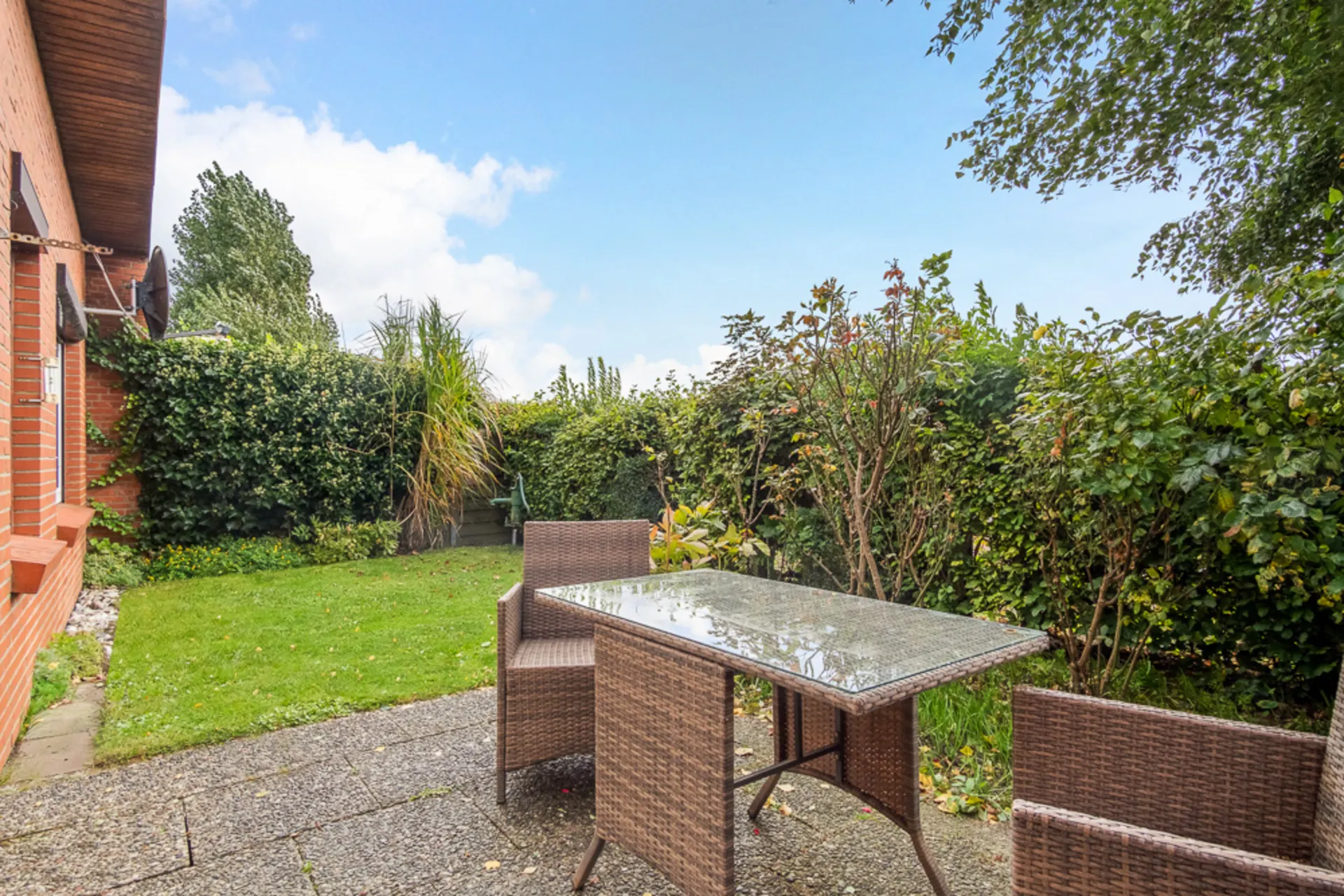 Gartenterrasse mit Glastisch und zwei Stühlen neben einem Backsteinhaus, umgeben von Hecken.