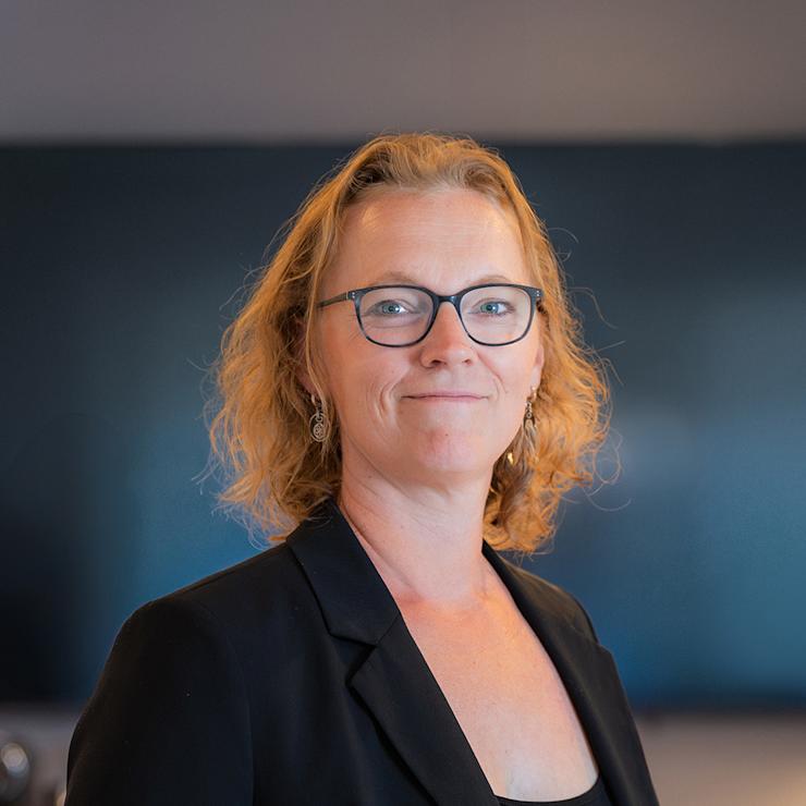 Eine Frau mit Brille und lockigen Haaren trägt ein blaues Sakko und steht vor einer beigen Wand.